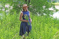 Die neue Projektleiterin der Genussregion Niederbayern steht im Uferbereich der Isar bei Landau. Sie trägt ein blaues Sommerkleid und einen Strohhut und hält einen Büschel Wildkräuter in den Händen.