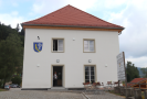 Blick auf den Eingang ins neue Bürgerhaus in Perasdorf. An der Außenfassade prangt das Gemeindewappen. Vorm Bürgerhaus stehen eine große Bautafel sowie mehrere Tische und Stühle.