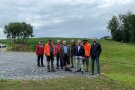 Auf der Kiesfläche vor dem neuen Rückhaltebecken stehen neun Herren. Im Hintergrund parken einige Autos. Daneben steht am Rande eines Maisfeldes ein Jäger-Hochsitz.