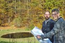 Das Foto zeigt zwei Herren, die den Baufortschritt am neuen Löschweiher in Ödhäuser begutachten. Die beiden halten einen großen Bauplan in ihren Händen. Im Hintergrund ist der mit Wasser gefüllte Löschweiher zu sehen. Am Waldrand dahinter erstrahlen die Blätter der Laubbäume in bunten Herbstfarben.