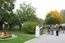 Links im Bild ist der neue Dorfbrunnen zu erkennen, der bei einem Festakt durch den Pfarrer in der Bildmitte offiziell eingeweiht wird. Rechts neben dem Geistlichen stehen mehrere Personen und lauschen seinen Worten. Im Hintergrund sind herbstlich anmutende Bäume und Sträucher zu sehen.