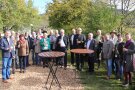 21 Frauen und Männer stehen rund um zwei Stehtische und stoßen mit frisch gepresstem Apfelsaft auf den gelungenen Schmankerlmarkt an. Im Hintergrund sind Streuobst- und Laubbäume sowie einige Marktstände zu sehen.