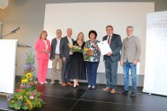 Drei Damen und vier Herren während eines Gruppenfotos im Rahmen der Preisverleihung auf der Bühne im Bürgerhaus Zeilarn.