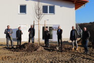 Vorm neuen Heizhaus der Nahwärmeversorgung in Hofham pflanzen sieben Männer einen Lindenbaum ein. Vier Herren halten einen Spaten in der Hand.