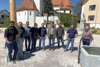 Neun Herren stehen nebeneinander auf dem neu gepflasterten Dorfplatz in Degernbach. Im Bild links befindet sich das Maibaumfundament samt Maibaum und am rechten Bildrand der neue Dorfbrunnen. Im Hintergrund sind die Kirche und das restaurierte Kriegerdenkmal zu sehen.