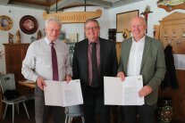 Drei Herren vollziehen die offizielle Übergabe einer Abschlussurkunde