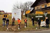 Acht Personen mit Masken stehen mit Spaten auf einer unbefestigten Fläche vor der Bautafel zur nun beginnenden Baumaßnahme im Rahmen der Dorferneuerung Wolfakirchen.