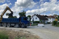 Ein blauer Schüttguttransporter wird von einem gelben Bagger mit Erdreich befüllt. Im Hintergrund ist der Ortseingang der Gemeinde Leibersdorf mit den ersten Wohnhäusern zu sehen.