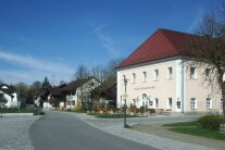 Eine neu asphaltierte Dorfstraße samt gepflasterten Gehsteig und Vorplatz. Im Hintergrund ist ein Gasthaus mit Biergarten zu sehen.