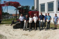 Sieben Männer und eine Frau stehen vor einem roten Bagger neben dem alten Schulhaus in Atzldorf. Fünf Personen halten symbolisch einen Spaten sowie einen weißen Bauhelm in den Händen. Im Hintergrund sind einige Wohnhäuser zu sehen.