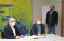 Eine Frau mit FFP2-Maske sitzt an einem Tisch und trägt sich ein Gästebuch ein. Daneben sitz ein Mann, auch im Hintergrund ist ein Mann, beide mit Maske zu sehen.