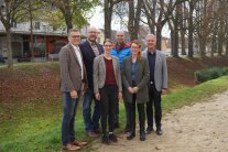 Vier Herren und zwei Damen stehen auf einem Kiesweg. Im Hintergrund ist ein begrünter Graben, Laubbäume und einige Geschäfte zu sehen.