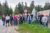 19 Personen, davon 3 Damen, sowie ein Schäferhund posieren nebeneinander für ein Gruppenfoto auf einem hergerichteten Kiesweg am Waldrand von Thyrnau. Hinter der Personengruppe ist ein Durchfahrts-Verbotsschild für PKW und Motorräder sowie ein ca. 3 Meter hoher Gedenkstein samt Erinnerungstafel zu sehen.