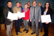 Sechs Herren und zwei Damen stehen leicht versetzt nebeneinander. Drei Personen halten eine gefaltete Urkunde in ihren Händen.