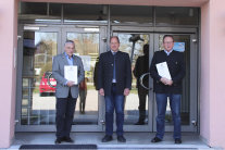 Drei Männer stehen vor einem Gebäude, zwei halten eine Urkunde in der Hand