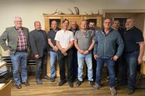 Zehn Herren stehen nebeneinander vor einem Holzschrank auf dem ein Geweih liegt.