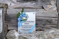 Eine rechteckige Alu-Plakette mit der Aufschrift „Auszeichnung 2023 Dorferneuerung & Baukultur – Ländliche Entwicklung in Niederbayern“. Die Kanten verzieren blau-weiße Banderolen.