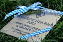 Eine rechteckige Alu-Plakette mit der Aufschrift „Auszeichnung 2023 Dorferneuerung & Baukultur – Ländliche Entwicklung in Niederbayern“ liegt im Gras. Die Kanten verzieren blau-weiße Banderolen.