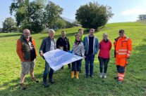 8 Personen stehen auf einer Wiese und halten einen sehr großen Plan gemeinsam in der Hand.