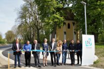 Neun Personen, davon sieben Herren und zwei Frauen durchtrennen mit ihren Scheren ein symbolisches blaues Band auf einem neu errichteten Geh- und Radweg. Rechts neben der Personengruppe ist eine graue Straßenlaterne zu sehen vor der ein Rollup mit dem Wappen der Stadt Neustadt an der Donau aufgestellt ist. Im Hintergrund ist die zurückgebaute B299 samt Geh- und Radweg sowie die Wittmann Kapelle zu sehen.