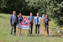 Drei Männer und zwei Frauen stehen auf einer Wiese rund um ein Baustellenschild mit der Aufschrift „Vermessung“ sowie einem Tachymeter herum. Im Hintergrund ist der Isardamm in Landau a.d.Isar und viele Laubbäume zu sehen.