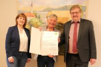 Zwei Herren und eine Frau stehen vor einem Gemälde der Ortsmitte von Kumreut. Der Mann in der Mitte hält einen Förderbescheid in seinen Händen.