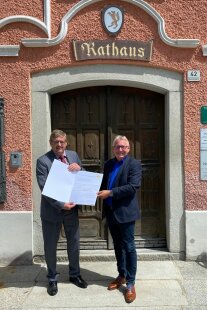 Zwei Herren im Anzug stehen vor der hölzernen Eingangstür zum Rathaus des Marktes Obernzell. An der Fassade über der Tür ist das Wappen und der Schriftzug „Rathaus“ zu sehen. Die beiden Männer halten gemeinsam eine Urkunde in ihren Händen.