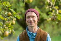 Das Foto zeigt einen grinsenden Mann mit blauen Augen und einer purpurfarbenen Strickmütze. Über seinen blauen Rollkragenpullover trägt er eine hellbraune Cord-Weste. Im Hintergrund sind herunterhängende Zweige eines Apfelbaumes zu sehen.