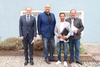 Vier Herren stehen vor zwei Fenstern an einer blauen Hauswand. Drei Männer halten Dokumente in ihren Händen. Am Boden hinter den Personen befindet sich ein betoniertes Beet mit grünen Bodendeckern.