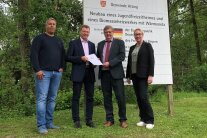 Drei Herren und eine Frau stehen auf einer Wiese vor der großen Bautafel zur einfachen Dorferneuerung Atting. Im Hintergrund sind mehrere Laubbäume zu sehen.