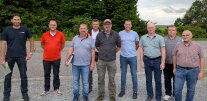 Neun Männer stehen auf einer asphaltierten Straße, im Hintergrund Sträucher und landwirtschaftliche Nutzfläche