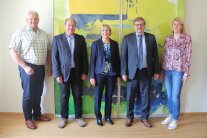 Beim Informationsbesuch der Regierung von Niederbayern versammeln sich drei Männer und zwei Frauen für ein Gruppenfoto. An der Wand hinter ihnen befindet sich eine teilbare Glaswand mit der künstlerisch gestalteten Silhouette von Landau a.d.Isar.