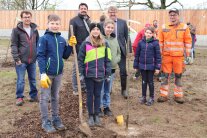 Vier Erwachsene und vier Kinder stehen rings um einen frisch gepflanzten Apfelbaum. Der Junge im Vordergrund hält einen Spaten in seiner linken Hand.