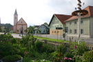 Im Vordergrund blüht es in einem mit Hanichlzaun umsäumten Bauerngarten mitten im Dorf von Eppenschlag, Lkr. Freyung-Grafenau. Im Hintergrund sind die Kirche, die Ortsstraße und Dorfhäuser zu sehen.