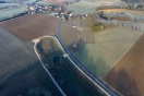 Vogelperspektive: Straße führt geschwungen in ein Dorf. Entlang der Straße Rückhaltebecken. Überlauf ermöglicht kontrollierten Wasserabfluss über den unscheinbaren Bach.