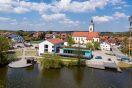 See Im Ortszentrum. Bühne im See mit Tribüne, dazwischen Seeweg. Daneben neues Rathausgebäude mit türkisenem Querbau. Dahinter bis zur Kirche Gemeinschaftsfläche für die Bürger.