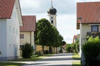 Eine Dorfstraße mit Grünstreifen und Laubbäumen führt auf die Dorfkirche zu.